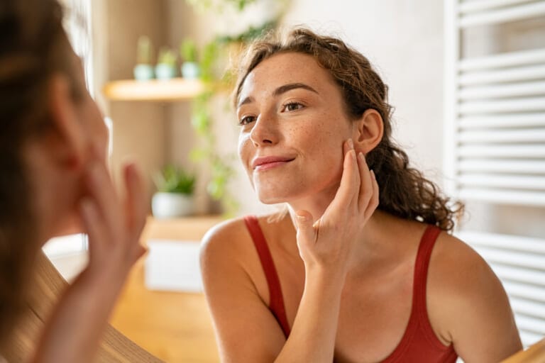 A woman looking at her face in the mirror and touching her skin.