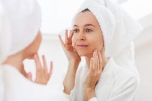 A middle-aged woman in a bathrobe and hair towel standing in front of a mirror, gently touching her face.