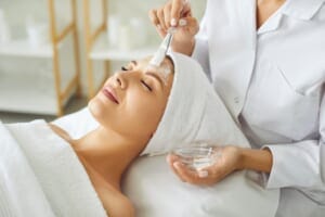 A moisturizing cream being applied with a brush to the face of a young, relaxed woman.