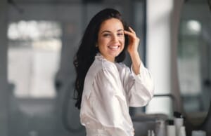 A young woman smiles radiantly while using skincare products in the bathroom.